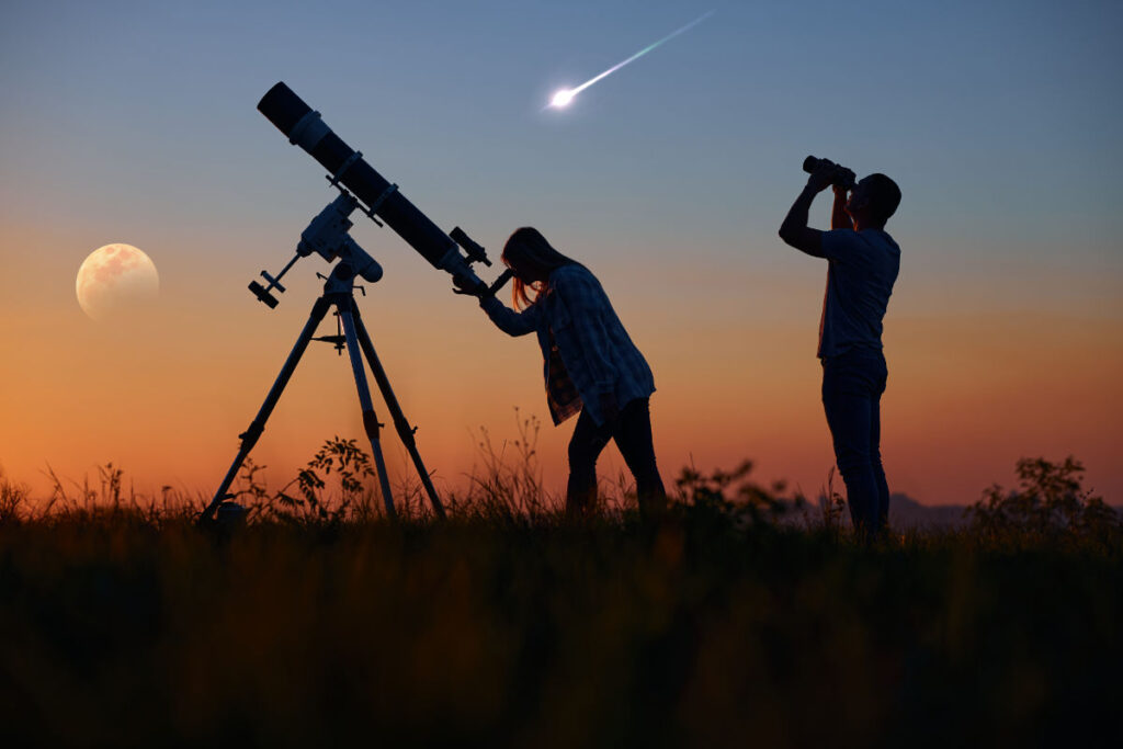 People stargazing with telescope and binoculars in open field with comet and moon in the sky